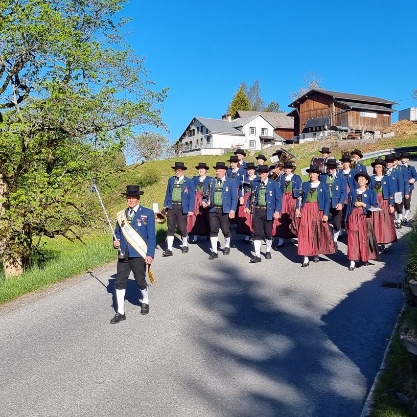 A group of people dressed in traditional costumes is marching down a road. They are accompanied by a musician playing a brass instrument. The setting is outdoors with houses and greenery in the background.