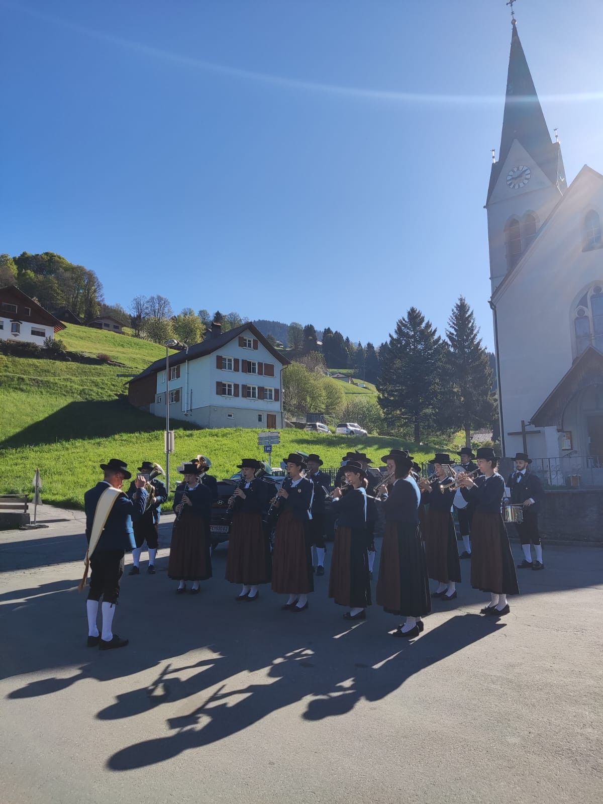 A band plays instruments in front of a church on a sunny day. Behind them is a grassy hill with houses.
