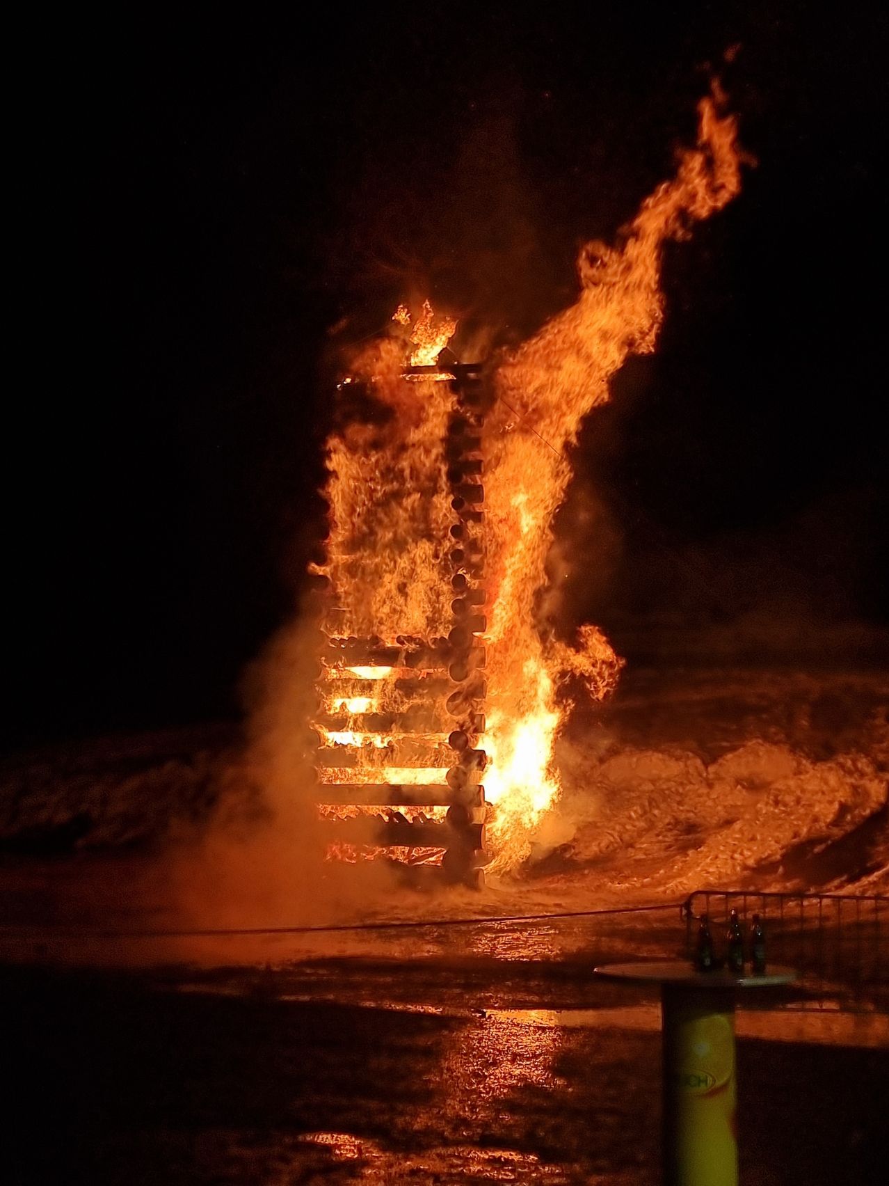 Ein großes Lagerfeuer brennt nachts hell, mit Flammen, die hoch reichen und Schatten auf den Boden werfen. Das Feuer ist von einem dunklen Bereich umgeben, möglicherweise einem Strand.