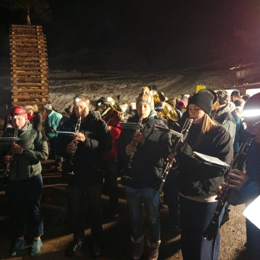 Eine Gruppe von Musikern, die Blasinstrumente halten, steht in einem Halbkreis in einem schwach beleuchteten Außenbereich und führt vielleicht eine Nachtaufführung durch.