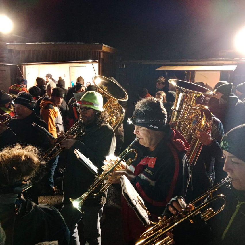 Eine Gruppe von Musikern tritt bei Nacht in einem überfüllten Außenbereich auf. Sie spielen verschiedene Instrumente, darunter Tubas und Trompeten. Die Menge wird von einem hellen Licht beleuchtet.