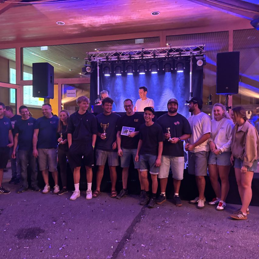 Eine Gruppe von Menschen steht auf einer Bühne, hält Pokale und posiert für ein Foto. Sie tragen Freizeitkleidung und einige tragen Brillen. Hinter ihnen ist eine Bühne mit schwarzem Hintergrund und einer Scheinwerfer. An den Seiten befinden sich Lautsprecher und eine Person steht vor der Bühne.
