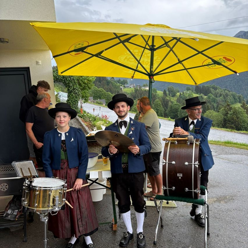 Drei Musiker stehen unter einem gelben Regenschirm. Die beiden Männer tragen traditionelle bayerische Uniformen. Eine Frau in der Mitte hält einen Trommelstock. Hinter ihnen stehen mehrere Personen, eine hält ein Bierglas. Im Hintergrund befinden sich Bäume und Berge.