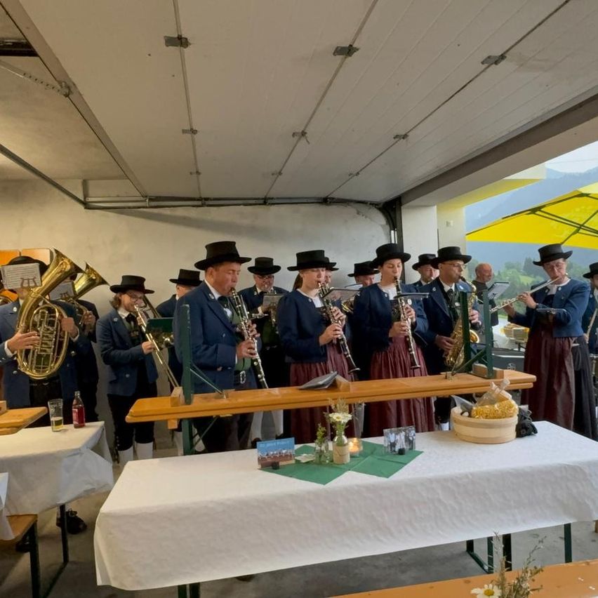 Eine Gruppe von Musikern, in traditioneller Kleidung gekleidet, steht hinter einem Tisch in einer Garage. Sie spielen verschiedene Instrumente, einige sitzend und andere stehend. Tische und Stühle umgeben sie.
