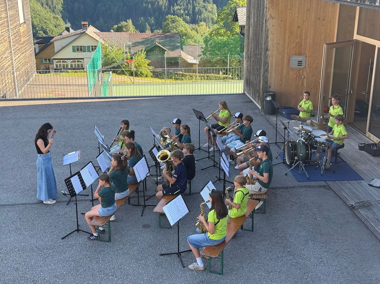 A group of young musicians sit on benches playing brass and woodwind instruments. They are outside with a grassy area and a building in the background.