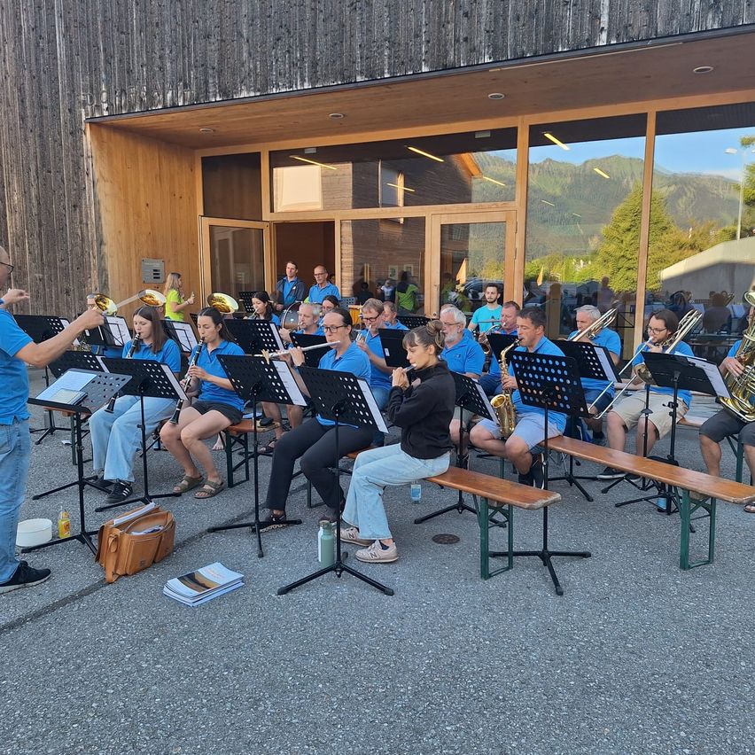A group of musicians are performing outside a building with large windows, showcasing a variety of instruments. The conductor stands before them with a baton.