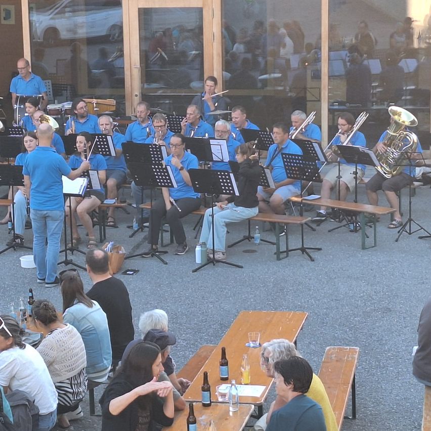 A band in blue shirts is performing outside. Spectators sit around tables, watching. Some have drinks, while others focus on the musicians. The setting is outdoors, with glass walls reflecting the scene.