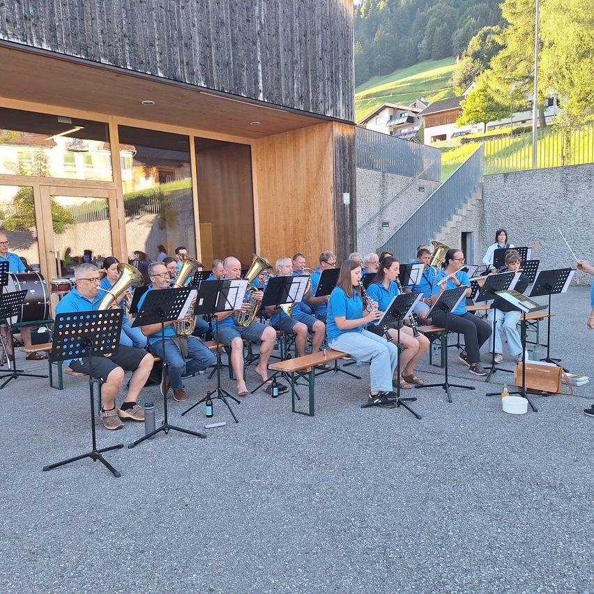 A brass band is playing in front of a building with glass doors. Members are seated on benches, holding instruments. The conductor stands to the right. Trees and houses are in the background.