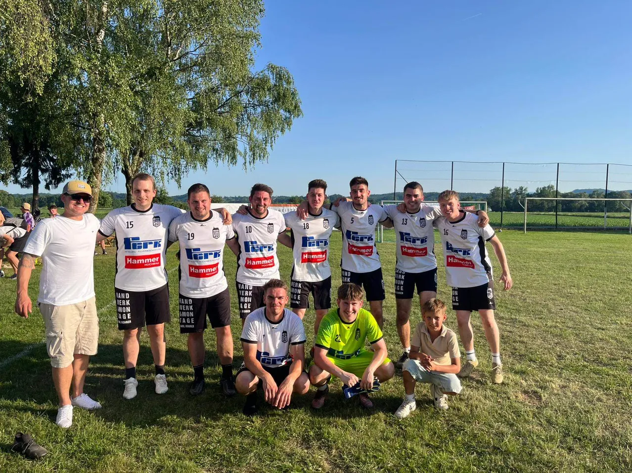 Eine Fußballmannschaft posiert für ein Gruppenfoto im Freien. Sie tragen weiße Trikots mit Nummern und schwarze Shorts. Zwei Jungen knien vorne. Dahinter steht ein Baum, ein Zaun und ein klarer blauer Himmel.