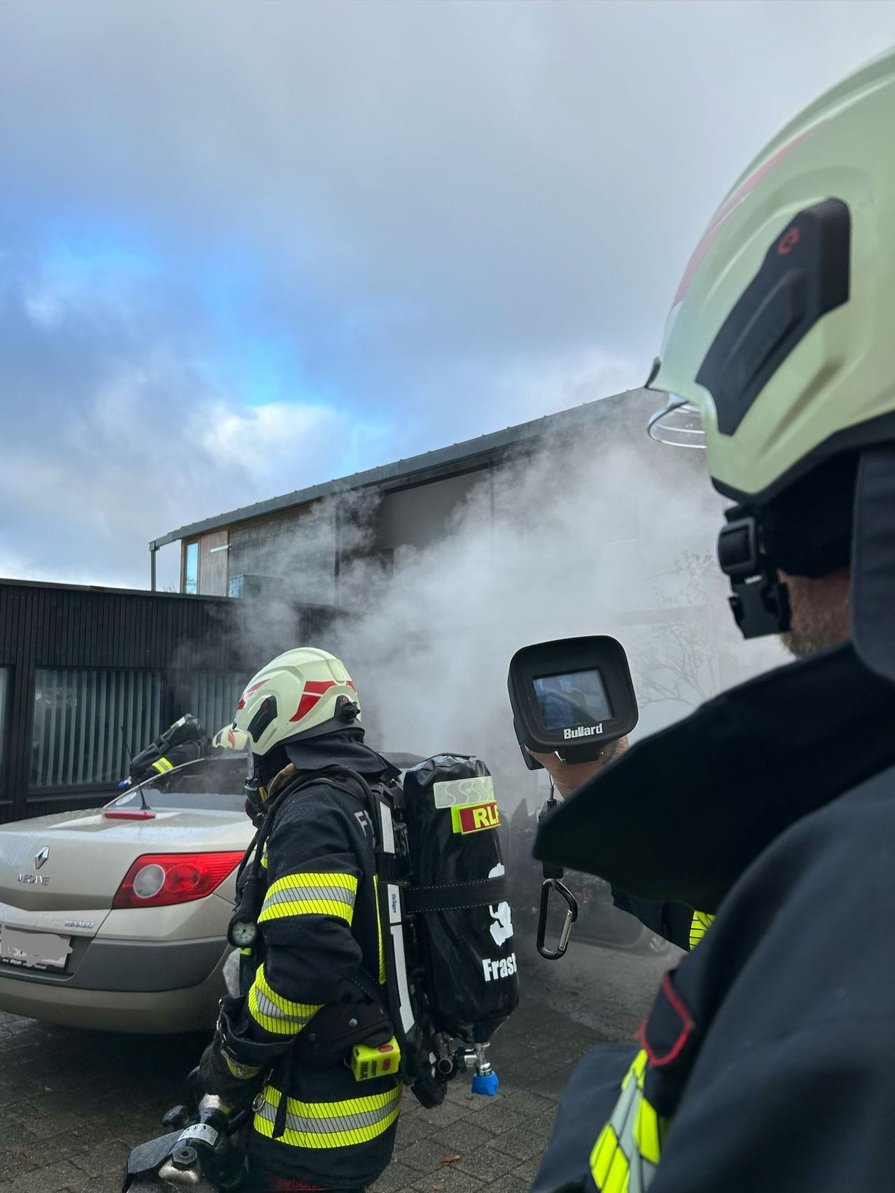 Feuerwehrleute in voller Ausrüstung stehen vor einem Gebäude, aus dem Rauch aufsteigt. Einer hält ein Gerät, möglicherweise eine Wärmebildkamera. Ein Auto ist in der Nähe geparkt.