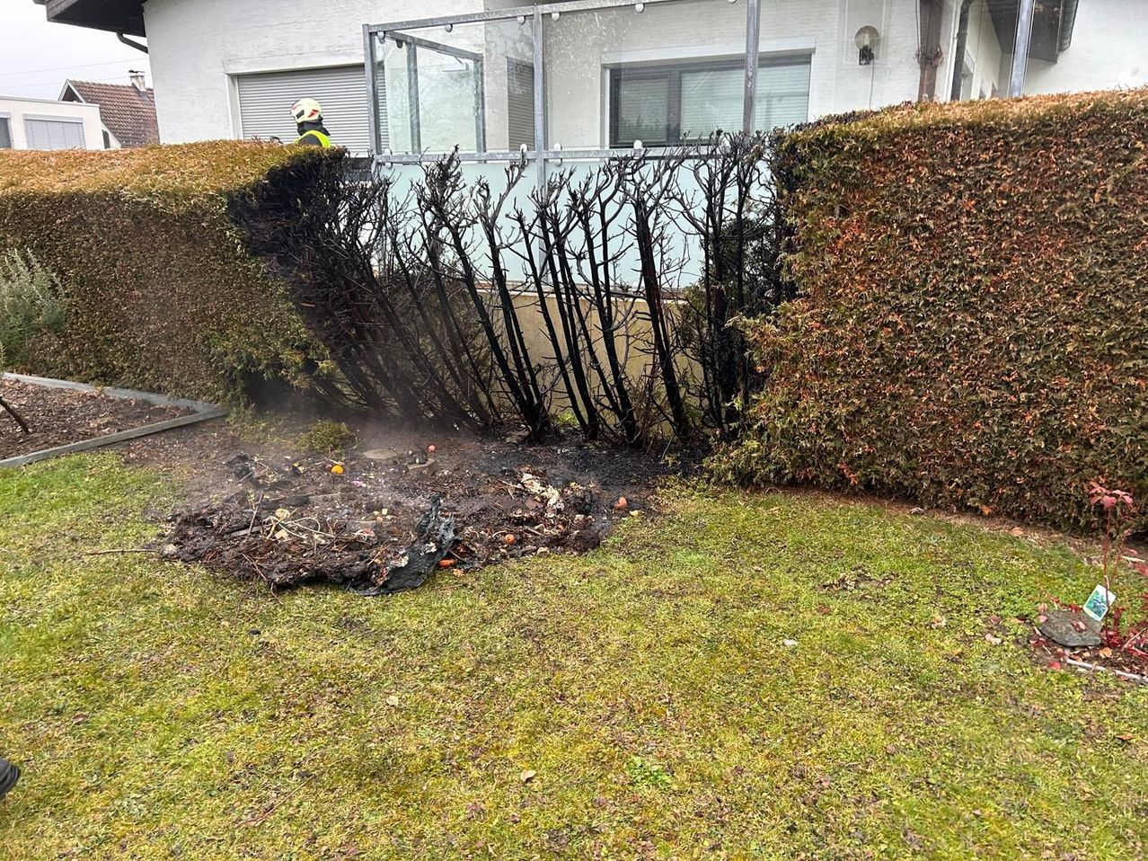 Ein Feuerwehrmann in gelber Jacke und Helm inspiziert einen verbrannten Busch im Garten eines Hauses mit Glasfenstern und Balkon.