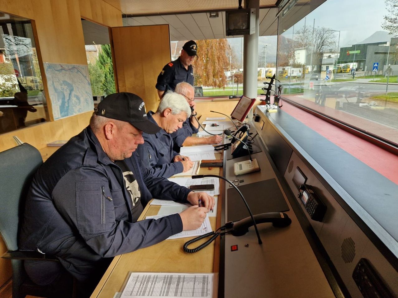 Three men in uniform sit at a desk in a room with large windows. They are looking at papers and one has a phone. A map is on the wall.