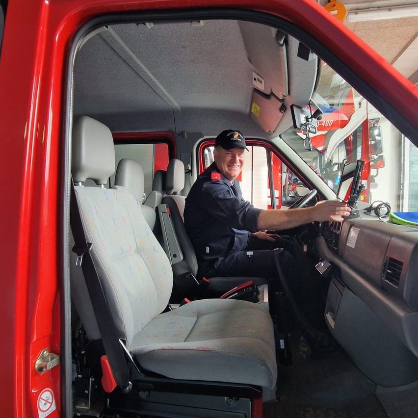 A firefighter is sitting in the driver's seat of a fire truck, smiling and holding the steering wheel. The interior of the truck is visible, with a red door, gray seats, and various control panels.