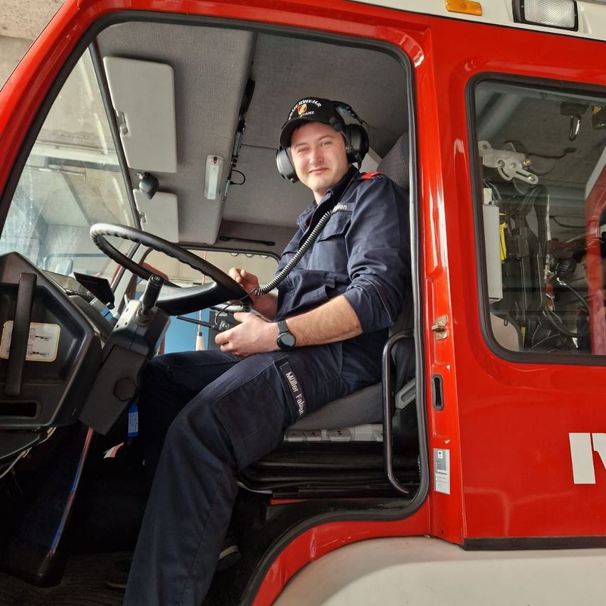 A firefighter sits in the driver's seat of a fire truck with the door open. He wears a watch and headphones, holding a tool in his hand.