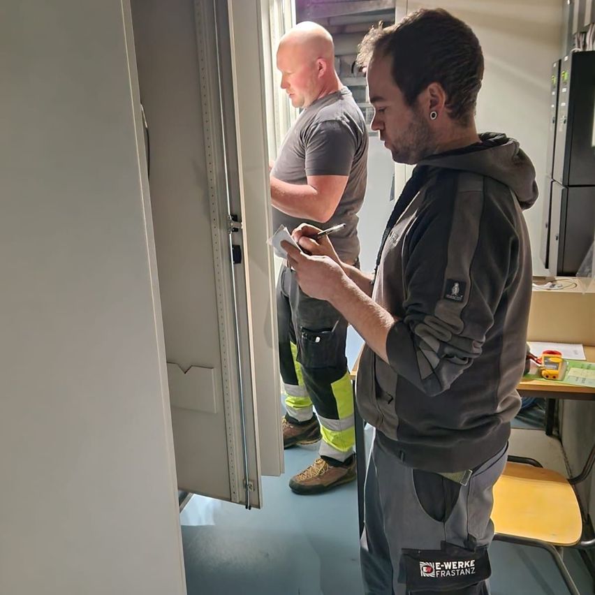 Two men in work attire are inside a room. One man is writing on a paper with a pen. The other man is standing behind him. They are near an open door.