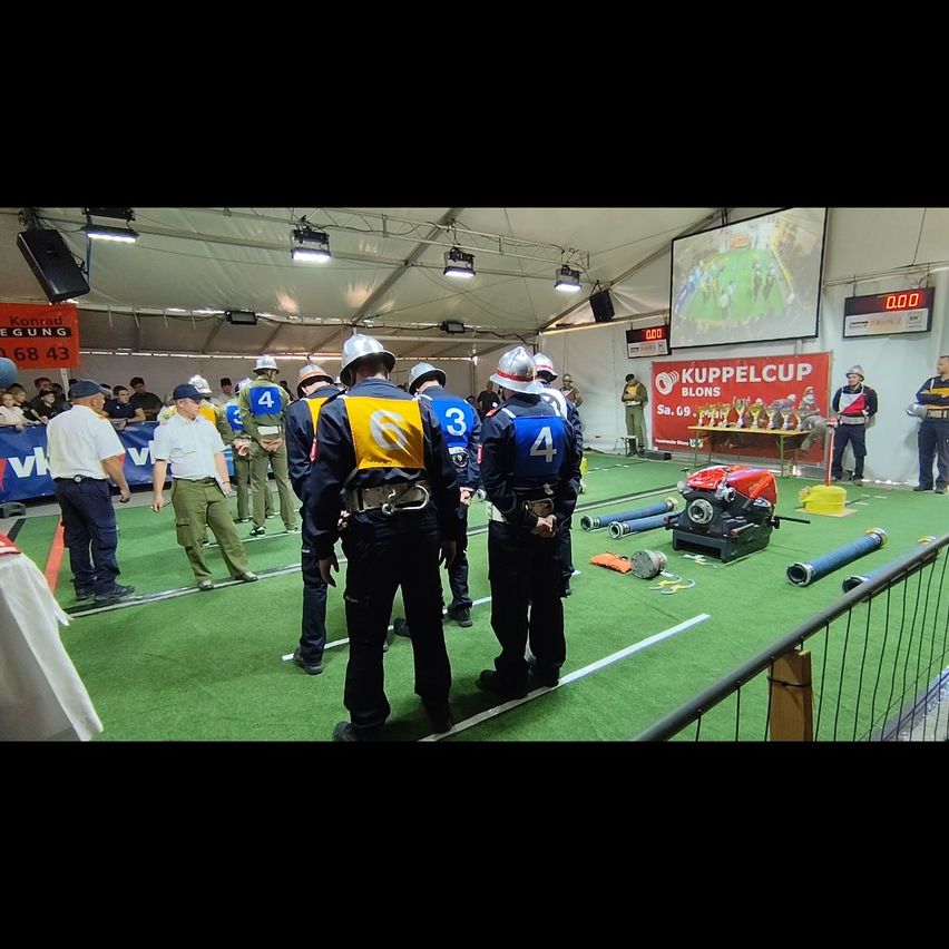 Teams of players are gathered in an indoor sports area, wearing uniforms and helmets. The ground is green, and a red banner with white text reads Kuppelcup.