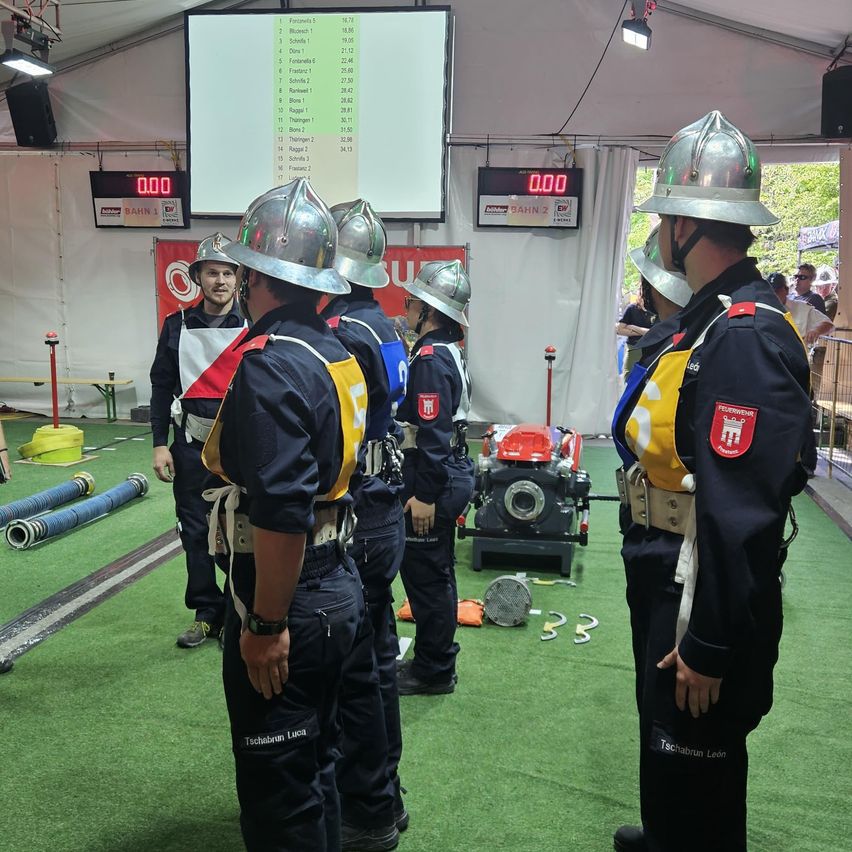 Firefighters in uniforms stand in a tent with a digital scoreboard. They are preparing for a competition. Equipment and machinery are on the floor.