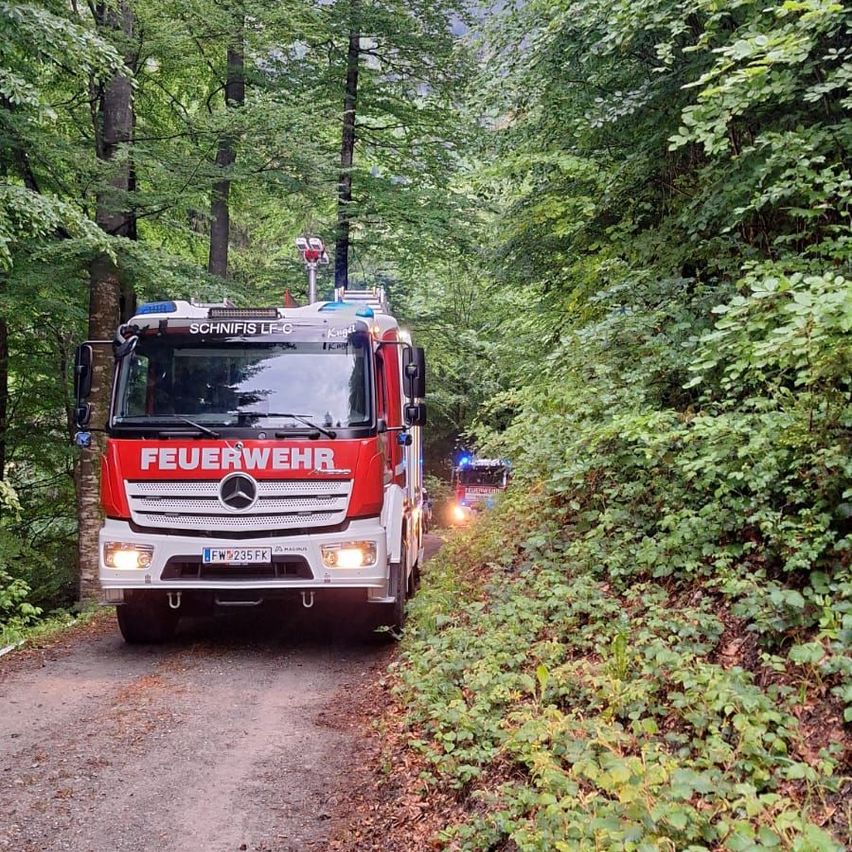 A red fire truck drives on a dirt road through a dense forest, with a police vehicle following behind. The fire truck has 'FEUERWEHR' written on it.