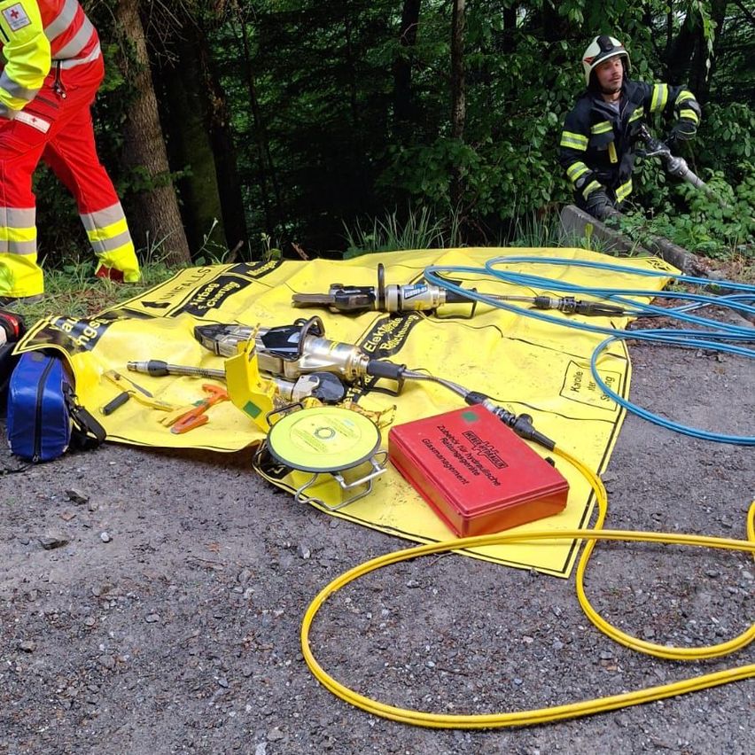 Two individuals in protective gear are in a wooded area. One is standing near a tree, and the other is crouched on the ground, holding equipment. Tools and a red box are on a yellow mat.