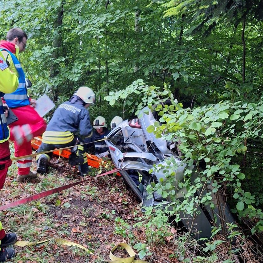 Emergency workers in helmets assist injured individuals near a wrecked car in a forest.