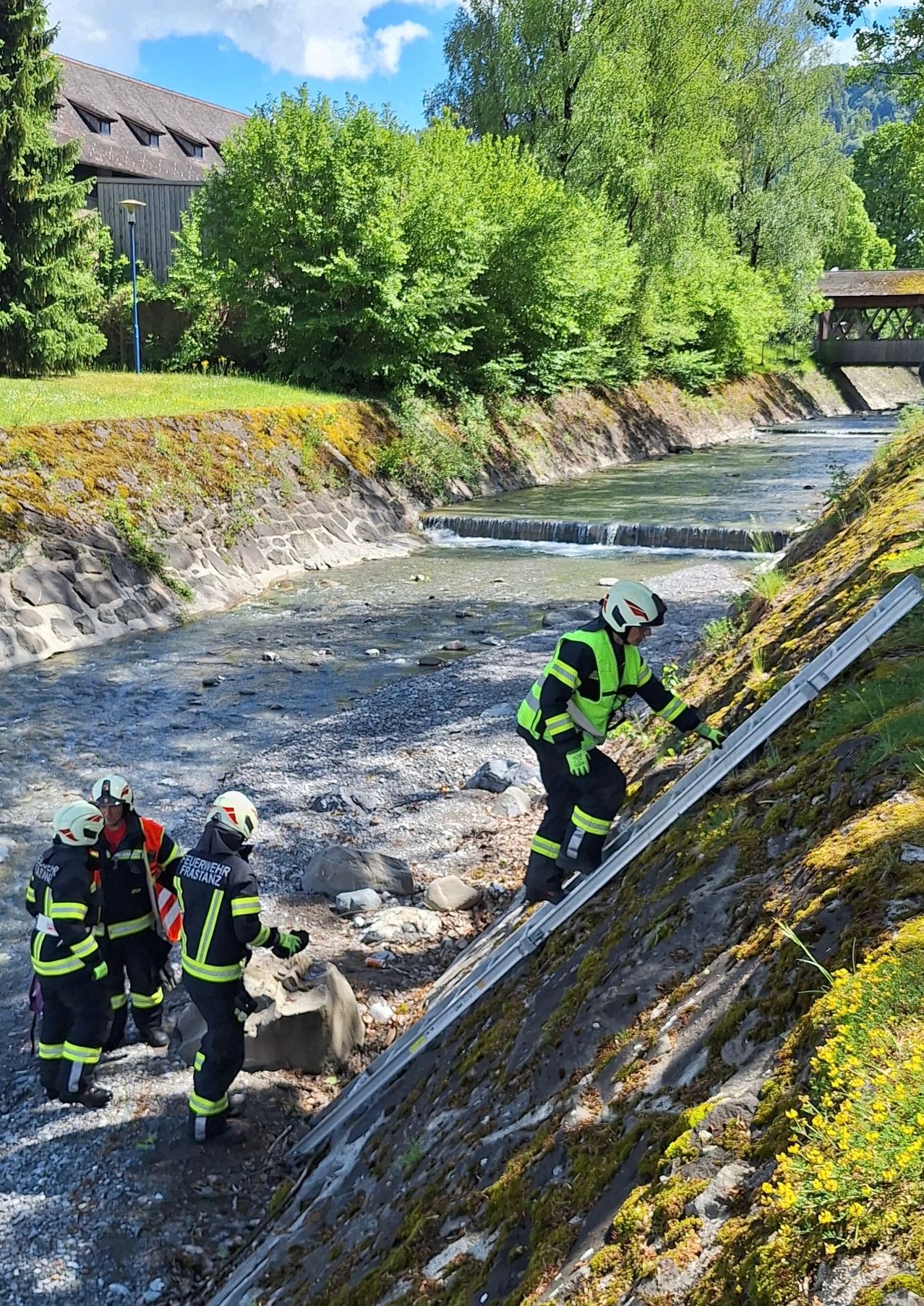 Firefighters are at the riverbank, one is climbing a ladder. The water flows through the rocks. Trees and grass are nearby.