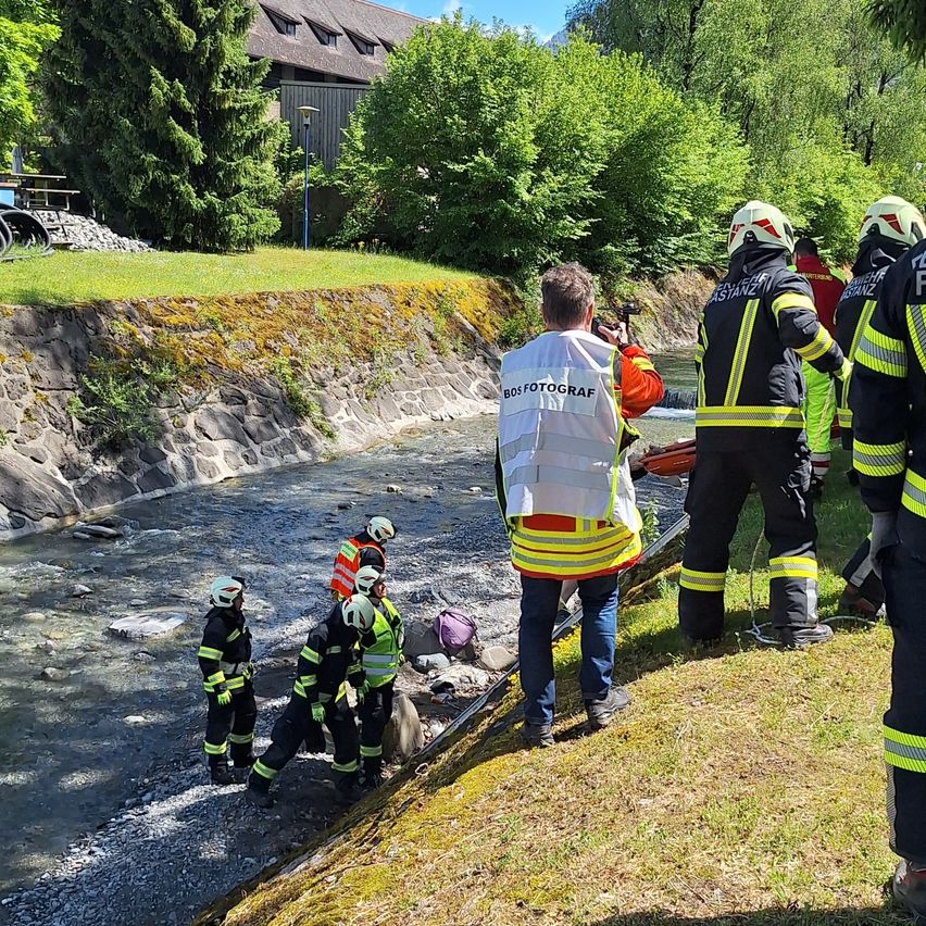 Firefighters in yellow and black uniforms are standing by a river, with one holding a camera and another looking at the water.