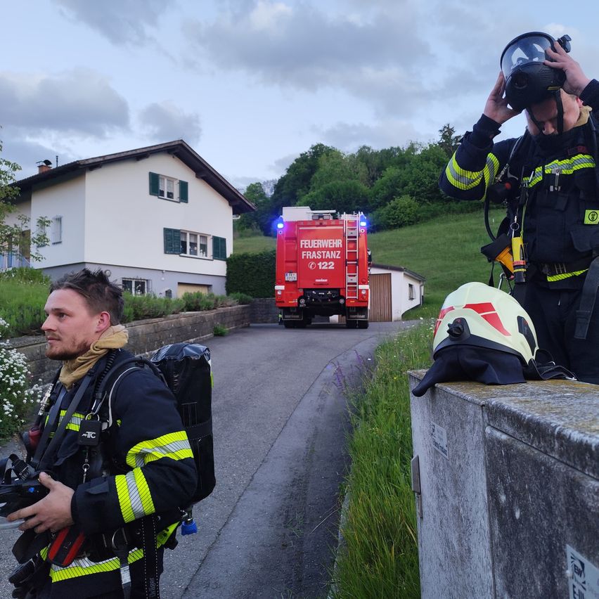 Two firefighters in full gear are standing on a street. One is holding a camera, while the other is wearing a gas mask. Behind them is a red fire truck.