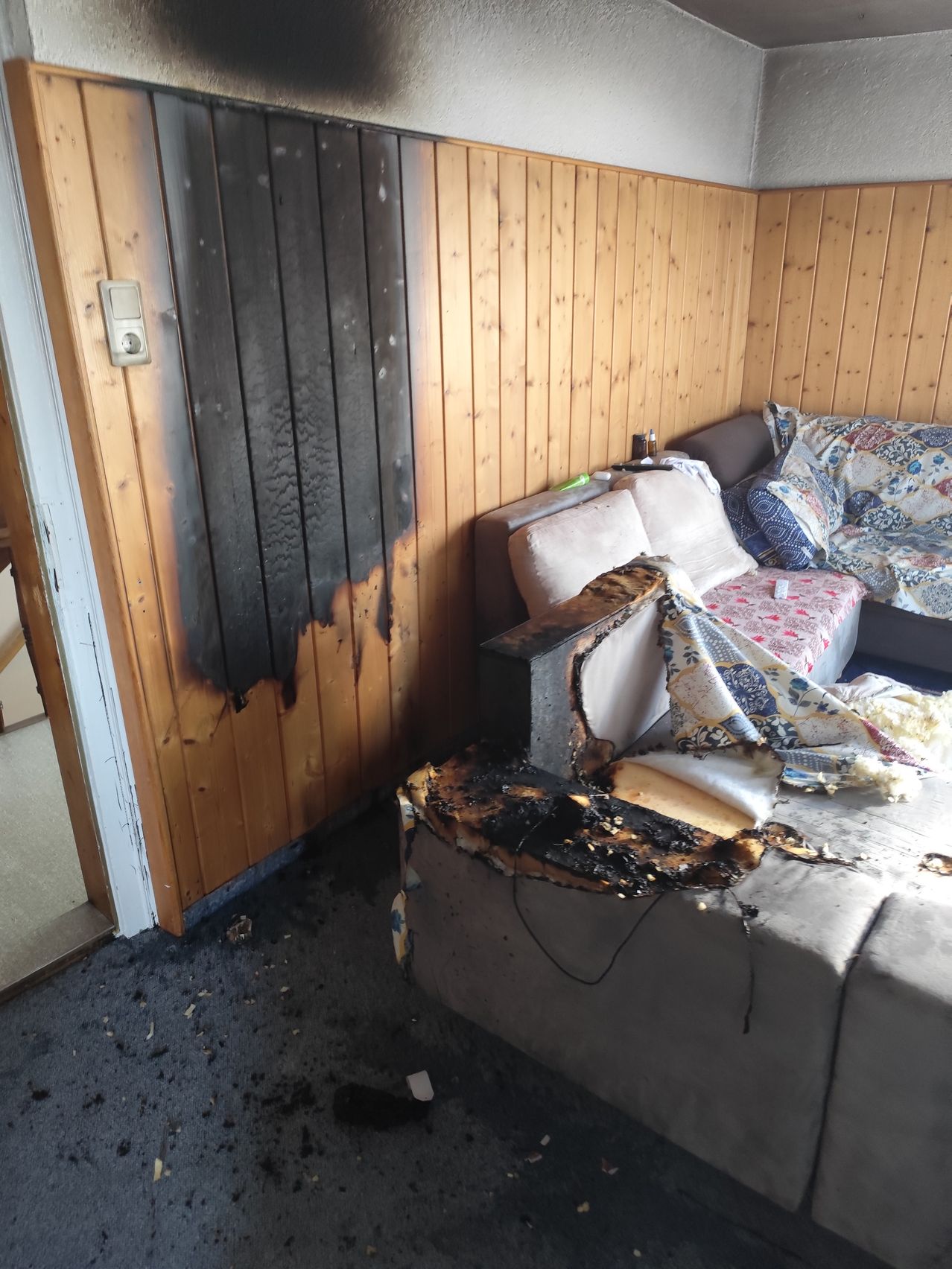 Ein Wohnzimmer mit einer verbrannten Holzwand und einem beschädigten Sofa. Das Sofa ist mit einer Decke und Kissen bedeckt.