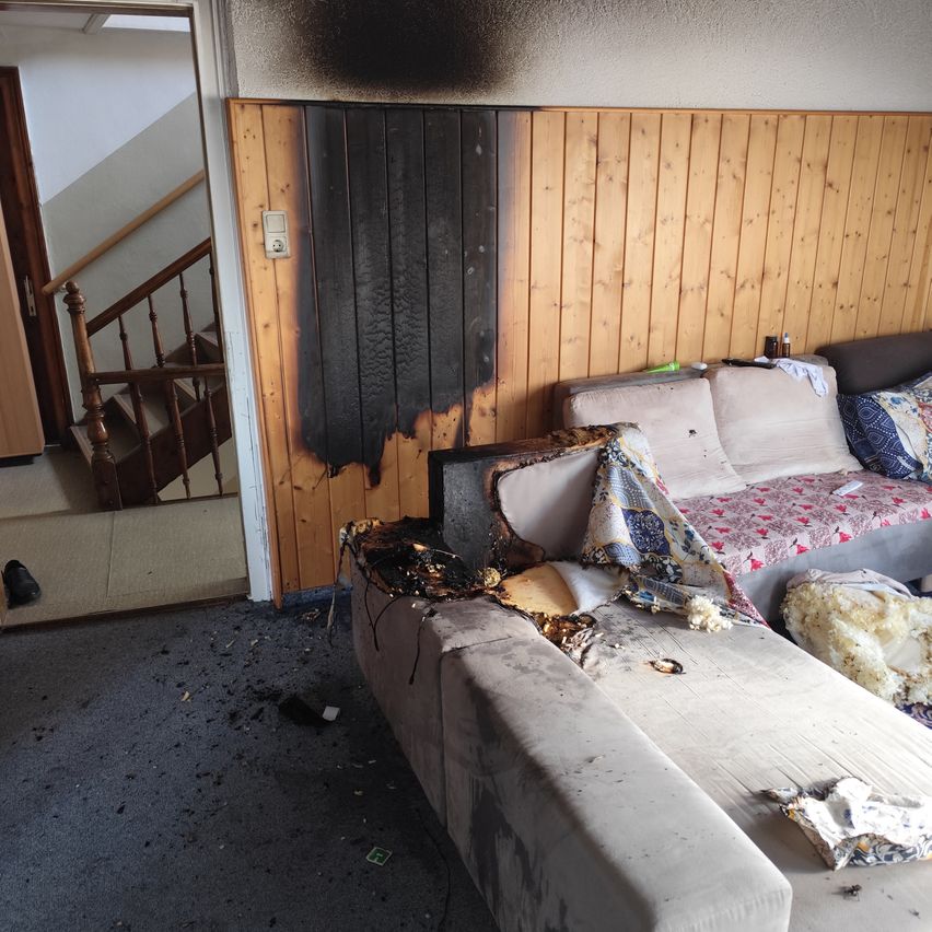 Ein Wohnzimmer mit einer verbrannten Holzwand und einem beschädigten Sofa. Der Raum hat eine Holztreppe und eine Tür, die zu einem anderen Raum führt.