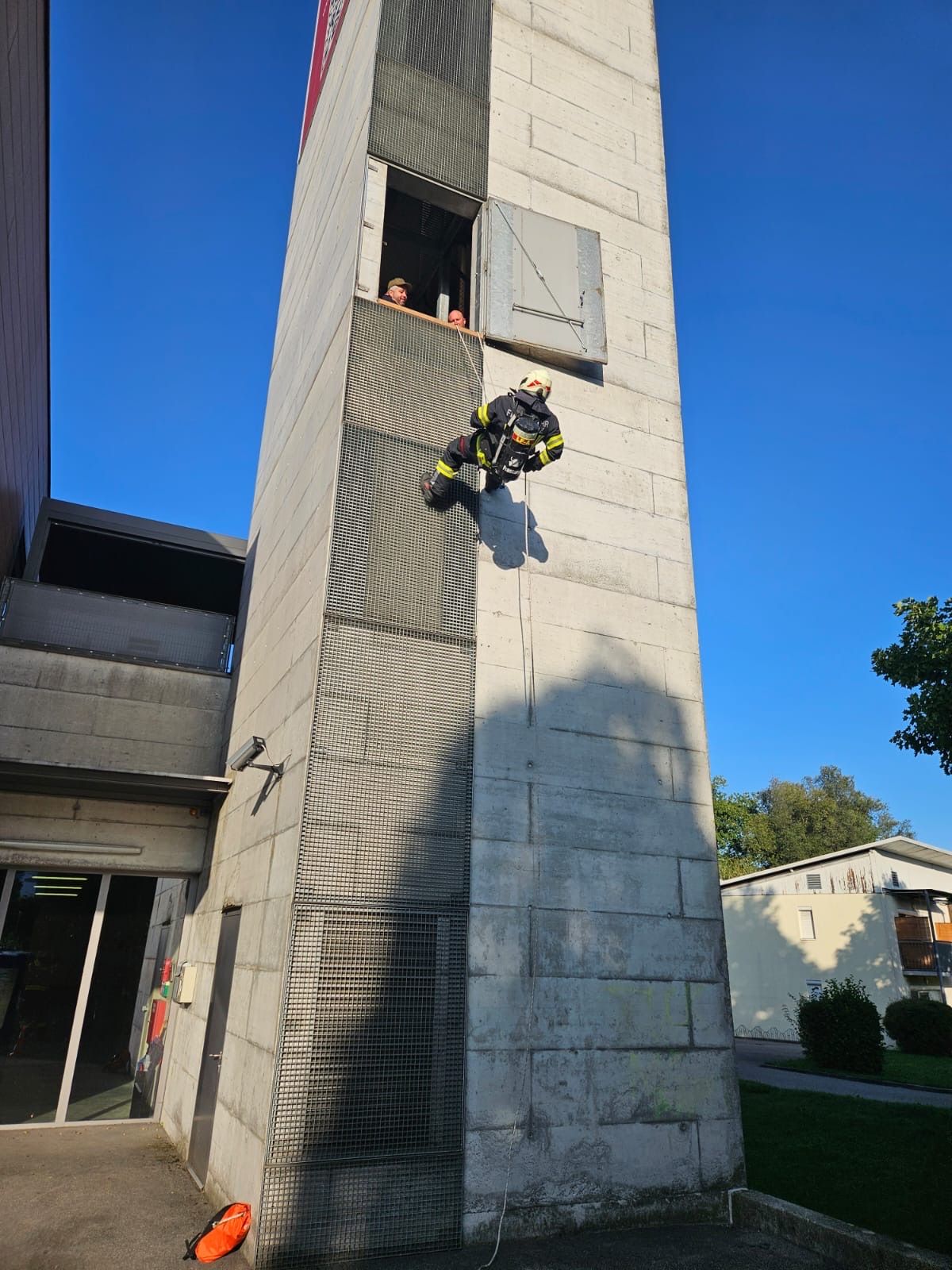 Ein Feuerwehrmann klettert mit einem Seil an einem Betongebäude hoch, während zwei Personen aus einem Fenster zusehen.