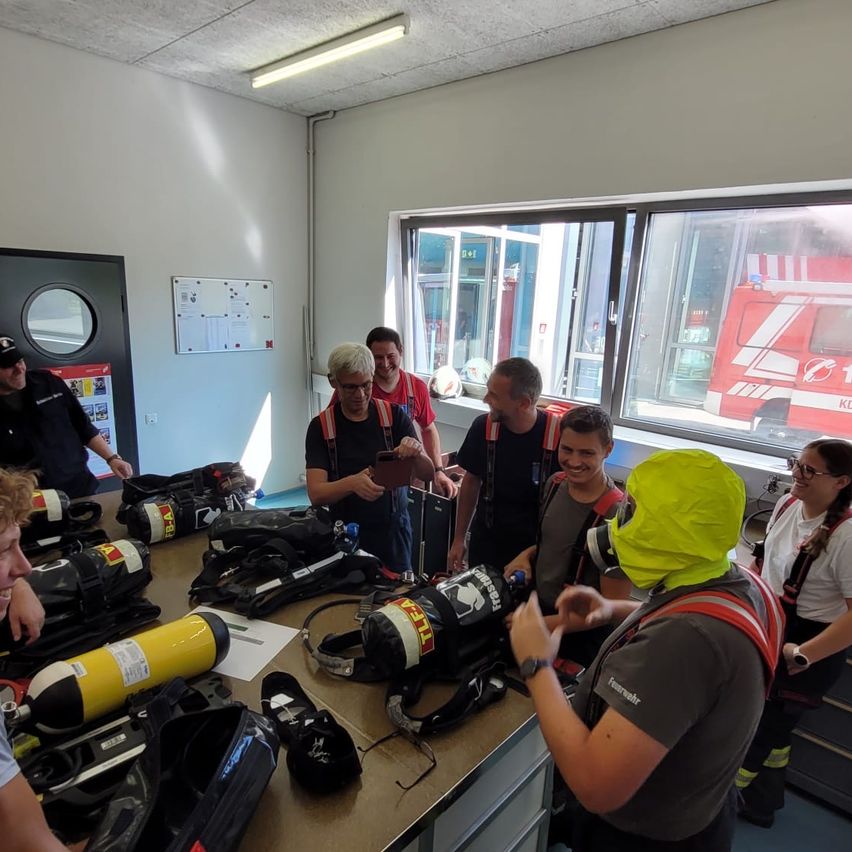 Eine Gruppe von Menschen in einem Raum mit Feuerschutzausrüstung, einschließlich Sauerstoffflaschen und Feuerlöschern, auf einem Tisch. Ein Mann in einem gelben Regenmantel spricht mit einer anderen Person.