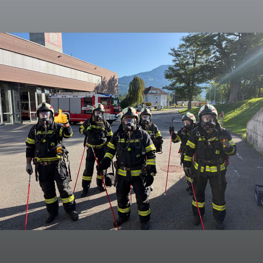 Eine Gruppe von Feuerwehrleuten in voller Ausrüstung, die in einer Reihe stehen und rote Stangen halten. Sie stehen vor einem Gebäude, vor dem ein Feuerwehrwagen geparkt ist.