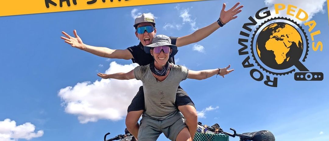 Zwei Radfahrer stehen mit ihren Fahrrädern auf der Straße, einer sitzt auf den Schultern des anderen. Das Poster bewirbt eine Radveranstaltung von Kapstadt nach Wien. Es erwähnt den Termin 26. Februar 2026 und den Ort Stadtsaal Z2000 Stockerau.