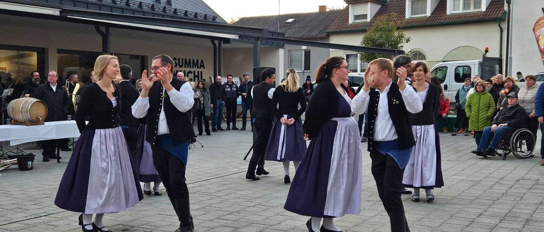 Menschen in traditioneller Kleidung tanzen vor einem Gebäude mit dem Wort 'Summa'. Dahinter stehen mehrere Personen in der Nähe eines schwarzen Objekts, möglicherweise eines Lautsprechers. Die Umgebung scheint ein Outdoor-Event zu sein.