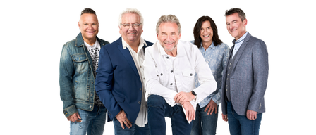 Fünf Menschen stehen lächelnd zusammen. Ein Mann sitzt auf einem blauen Hocker. Zwei Frauen und drei Männer, alle in Jeans. Sie sind lässig gekleidet. Der Hintergrund ist weiß.