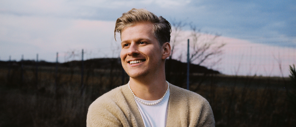Ein junger Mann mit blonden Haaren, lächelnd, steht in einem Feld. Er trägt ein weißes T-Shirt und einen beigen Cardigan, dazu eine Perlenkette. Hinter ihm ein Feld mit trockenem Gras und einem Baum.