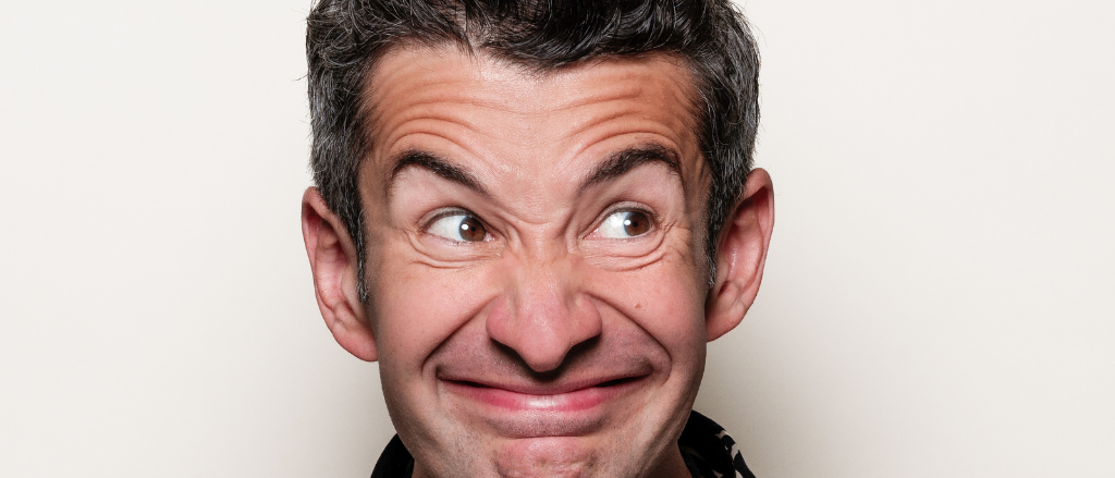 Ein Nahaufnahme eines lächelnden Mannes mit grauem Haar, der ein schwarz-weißes Hemd und eine schwarze Halskette trägt. Er hat Falten um die Augen und schaut nach oben.