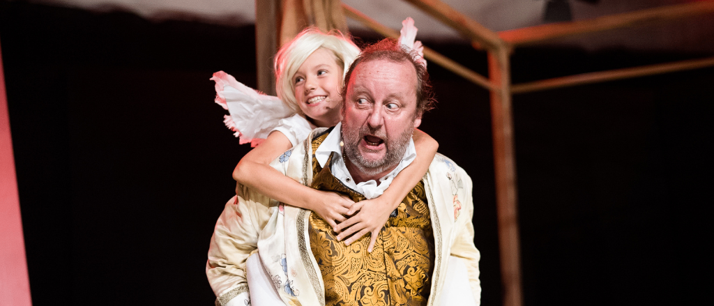 Ein Mann in einem aufwendigen Kostüm hält ein lächelndes Kind mit Flügeln auf dem Rücken auf der Bühne.