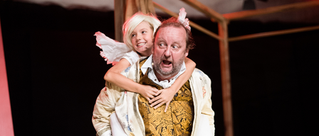 Ein Mann in einem aufwendigen Kostüm hält ein lächelndes Kind mit Flügeln auf dem Rücken auf der Bühne.