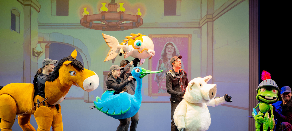 Eine Gruppe von Darstellern in Tierkostümen auf der Bühne, einer hält eine Fischpuppe, der andere eine Vogelpuppe. Dahinter zeigt eine Kulisse ein Schloss, eine Frau und eine Laterne.