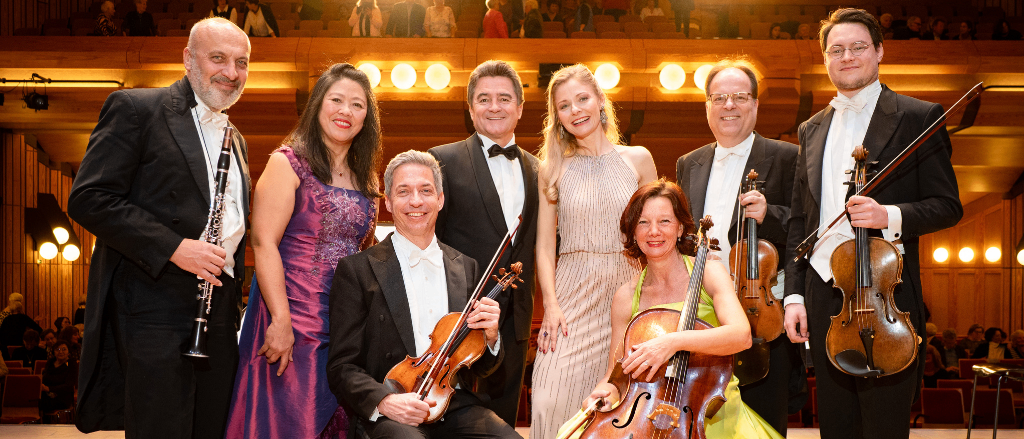 Eine Gruppe von Musikern in formeller Kleidung posiert für ein Foto auf einer Bühne mit Saiten- und Blasinstrumenten. Eine Frau in einem grünen Kleid hält ein Cello, während ein Mann in einem Anzug die Geige spielt.