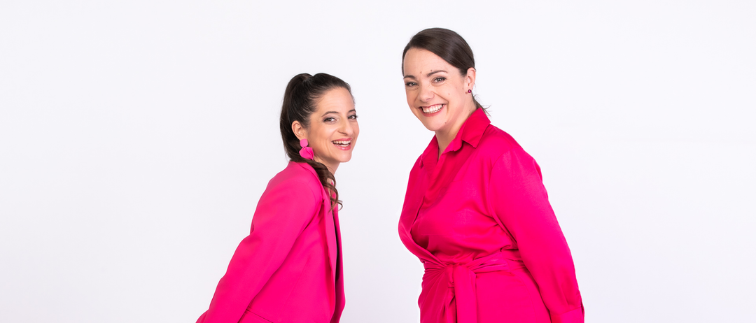 Zwei Frauen in rosa Outfits lächeln und posieren für ein Foto vor einem weißen Hintergrund. Die Frau links trägt einen rosa Blazer und eine Hose, während die Frau rechts ein rosa Kleid trägt.