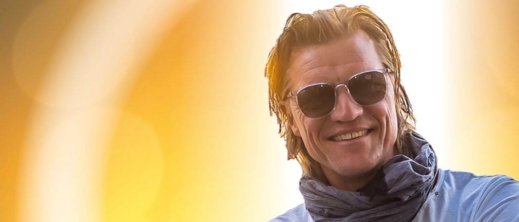 Ein Mann mit feuchten Haaren, der eine Sonnenbrille und einen Schal um den Hals trägt, lächelt breit. Er trägt ein weißes T-Shirt.