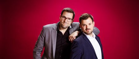 Zwei Männer stehen eng beieinander vor einem roten Hintergrund, beide in formeller Kleidung. Der Mann auf der linken Seite trägt eine Brille und hat seine Hand in der Tasche. Der Mann auf der rechten Seite hat seine Hand in der Tasche und seinen Arm um die Schulter des anderen Mannes gelegt.