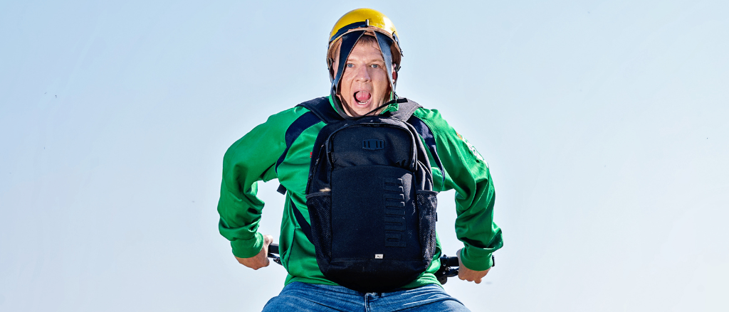 Ein Mann mit einem gelben Helm und einem grünen Jackett fährt mit einem Rucksack auf dem Rücken ein Fahrrad auf einem Feld.