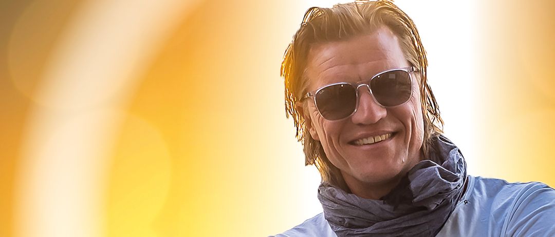 Ein Mann mit einem Schal um den Hals und Sonnenbrille auf den Augen lächelt und posiert mit verschränkten Armen.