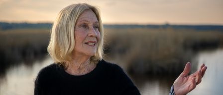 Eine ältere Frau mit kurzen blonden Haaren lächelt und gestikuliert mit ihrer Hand in der Nähe eines ruhigen Gewässers.