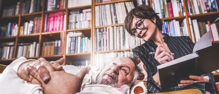 Ein älterer Mann liegt auf einer Couch und schaut eine Frau an, die ein Buch und einen Stift hält und in einer Bibliothek lächelt.