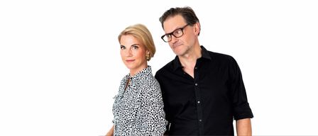 Ein Mann und eine Frau stehen eng beieinander und lächeln. Die Frau trägt ein schwarz-weißes Blusenkleid mit Punkten, der Mann ein schwarzes Hemd. Sie stehen vor einem schlichten weißen Hintergrund.