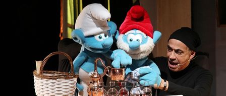 Zwei blaue Schlumpf-Puppen, eine mit einem Kochmütze und die andere mit einem roten Hut, stehen neben einem Mann in einem schwarzen Shirt. Sie sind auf einem Holztisch mit einem Korb und einem Kupferdestillationsapparat.